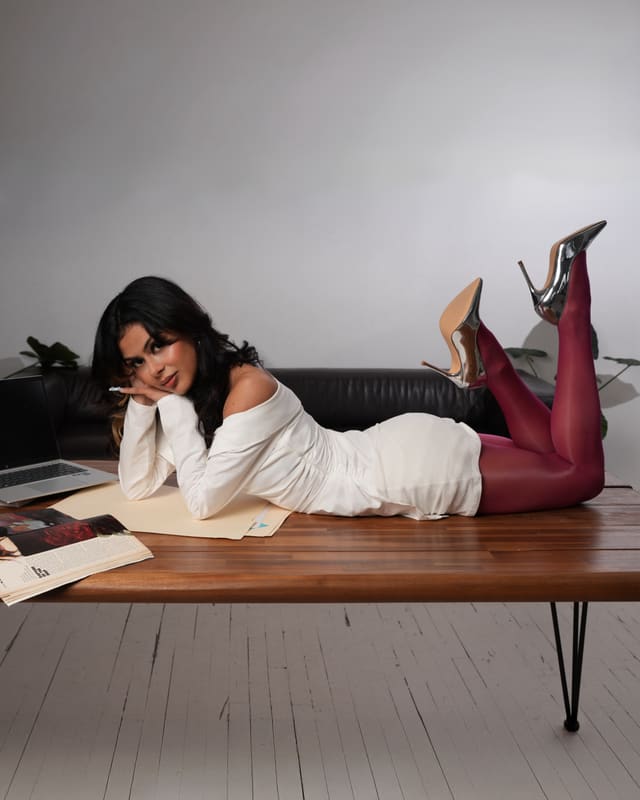 Lying across a wooden desk in a white dress and burgundy tights